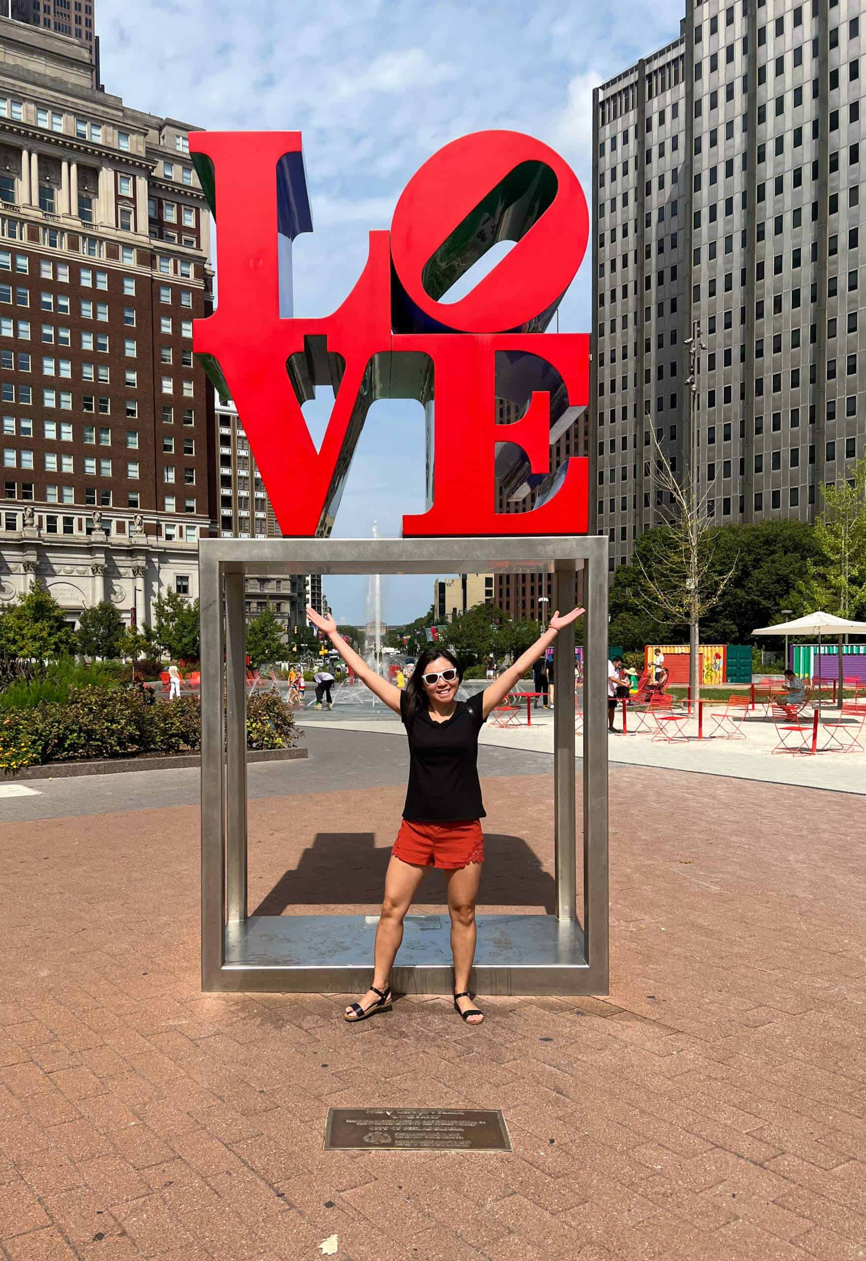 Standing in front of the LOVE sculpture in Love Park, Philadelphia. It’s a hot and bright summer day and the sculpture is book-cased by 2 historic Philadelphia buildings.