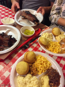 Two trays of BBQ and sides, including ribs, the dinosaur bone, mac and cheese, cornbread, and potato salad.