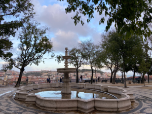 A fountain and some trees at Miradouro de São Pedro de Alcântara with tourists in the background taking pictures of the viewpoint of the city. One of my favorite places in this travel guide to Lisbon. 