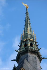 The spire at Mont Saint-Michel in France with a blue sky and a golden Saint-Michel on top.