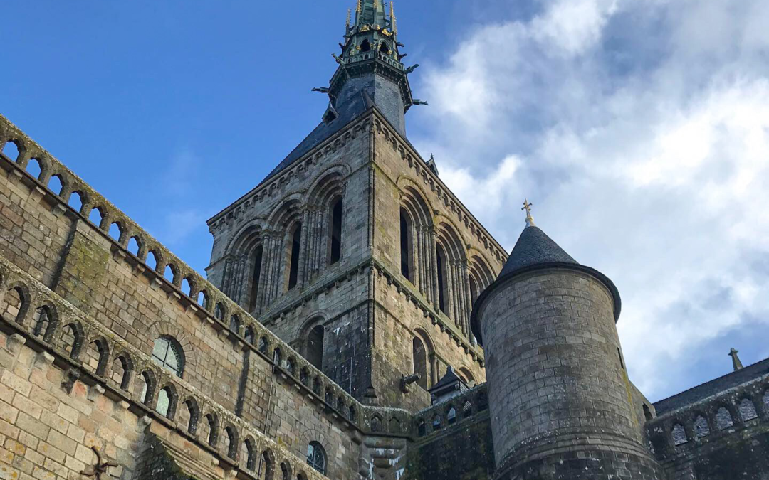 Looking up towards the tower and spire at Mont Saint-Michel in France with blue skies and some clouds.