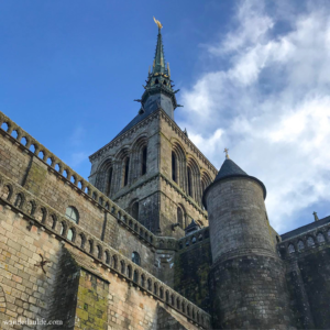 Looking up towards the tower and spire at Mont Saint-Michel in France with blue skies and some clouds.